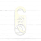A white door hanger with the text "THE WITCH IS IN" printed on it, along with a silhouette of a witch.