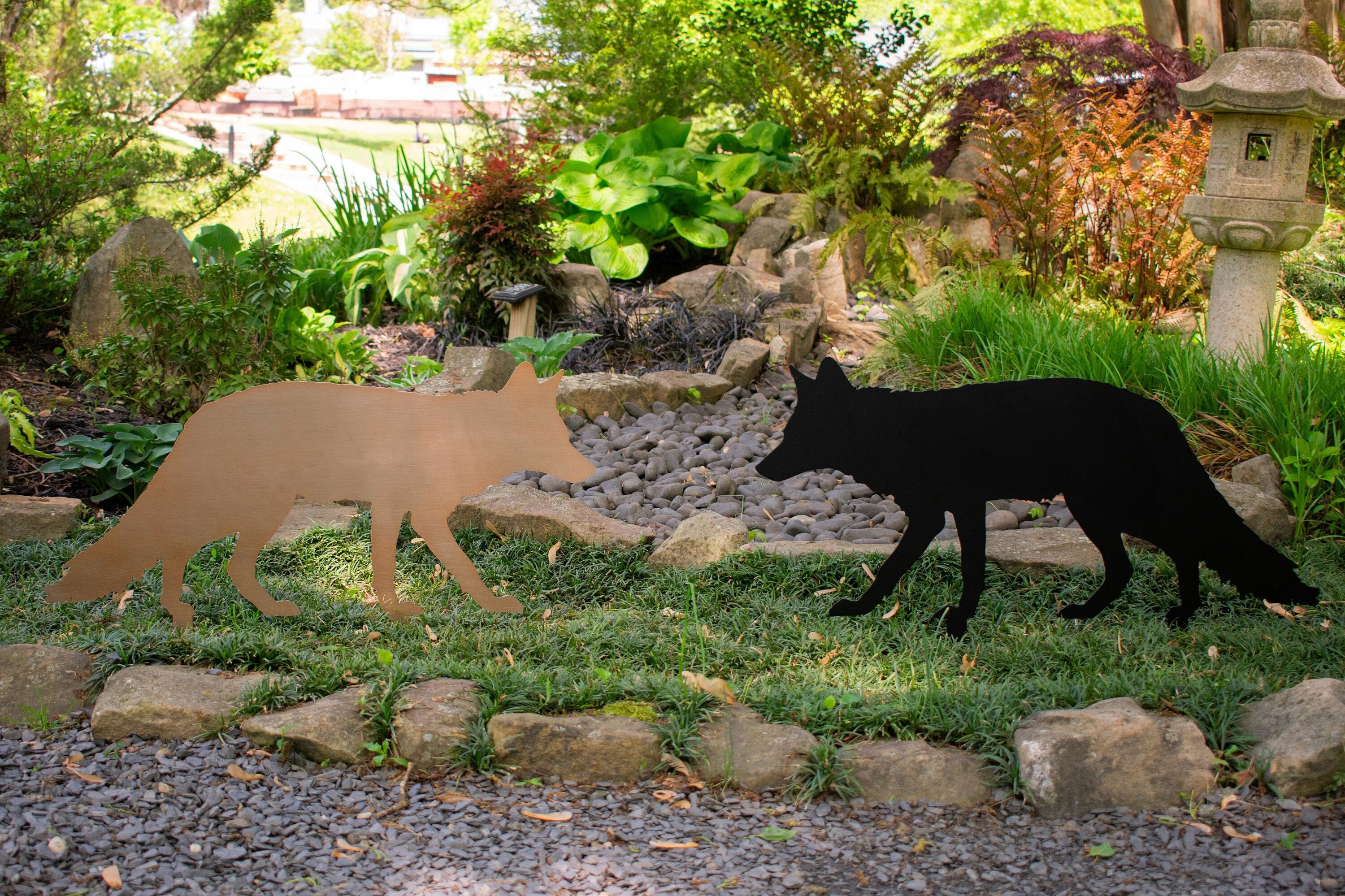 One copper fox yard stake and one black one. Facing towards one another with beautiful vegetation in the background