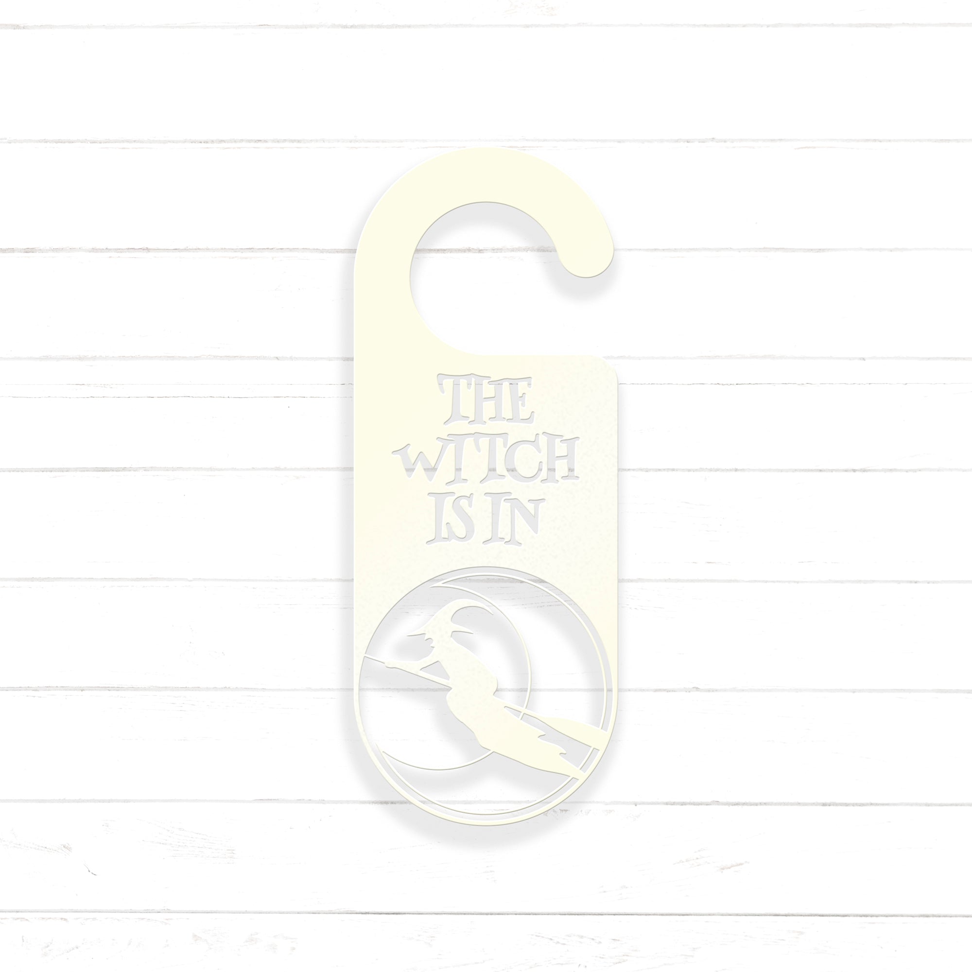 A white door hanger with the text "THE WITCH IS IN" printed on it, along with a silhouette of a witch.