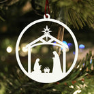 A white ornament hanging on a Christmas tree, depicting the nativity scene with the baby Jesus, Mary, and Joseph in a manger.
