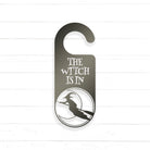 A black and white sign with the text "THE WITCH IS IN" and a silhouette of a witch on a broomstick.