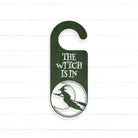 A green door hanger with the text "THE WITCH IS IN" printed on it, along with a silhouette of a witch riding a broomstick.