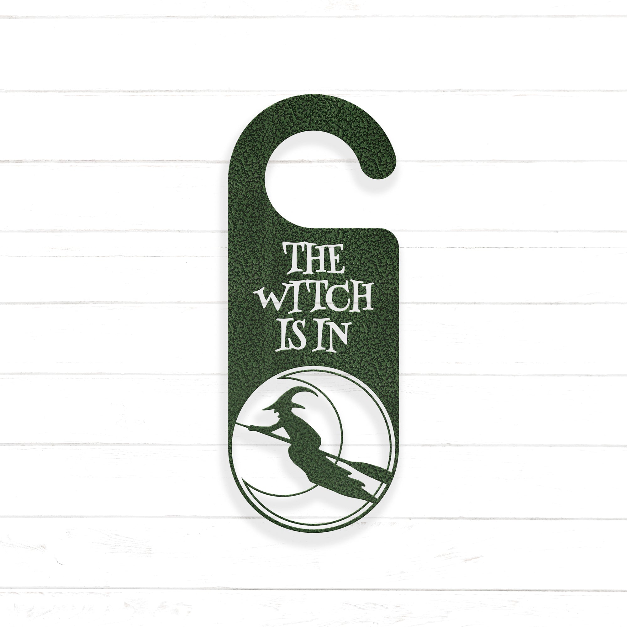 A green door hanger with the text "THE WITCH IS IN" printed on it, along with a silhouette of a witch riding a broomstick.