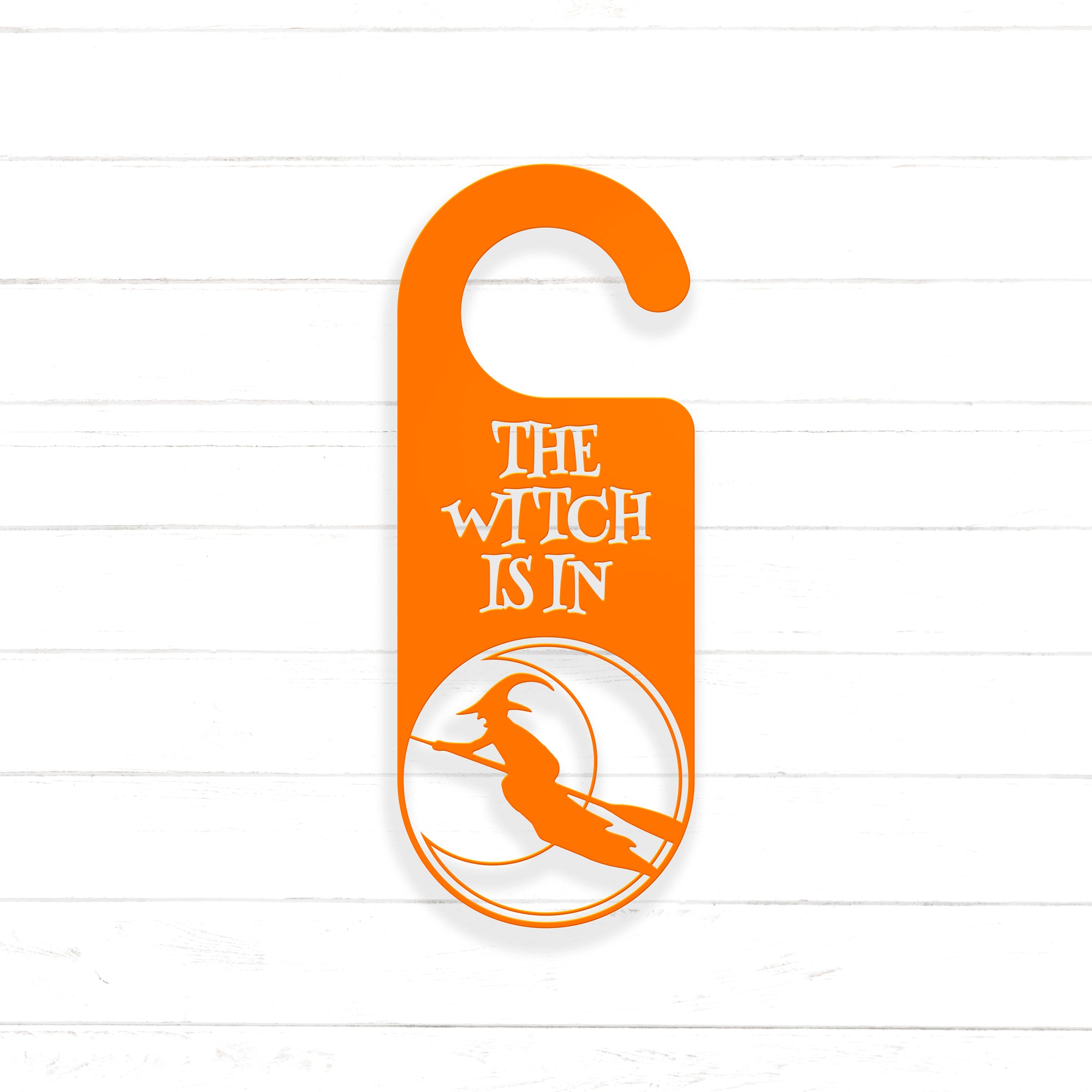An orange door hanger with the text "THE WITCH IS IN" printed on it, along with a silhouette of a witch riding a broomstick.